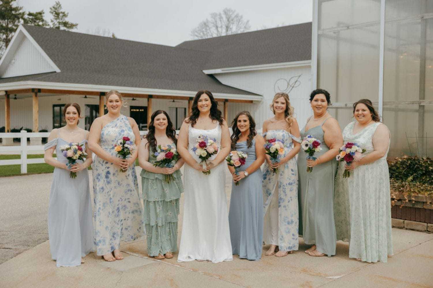 Bridal party with bouquets in pastel dresses standing outdoors near a white building.
