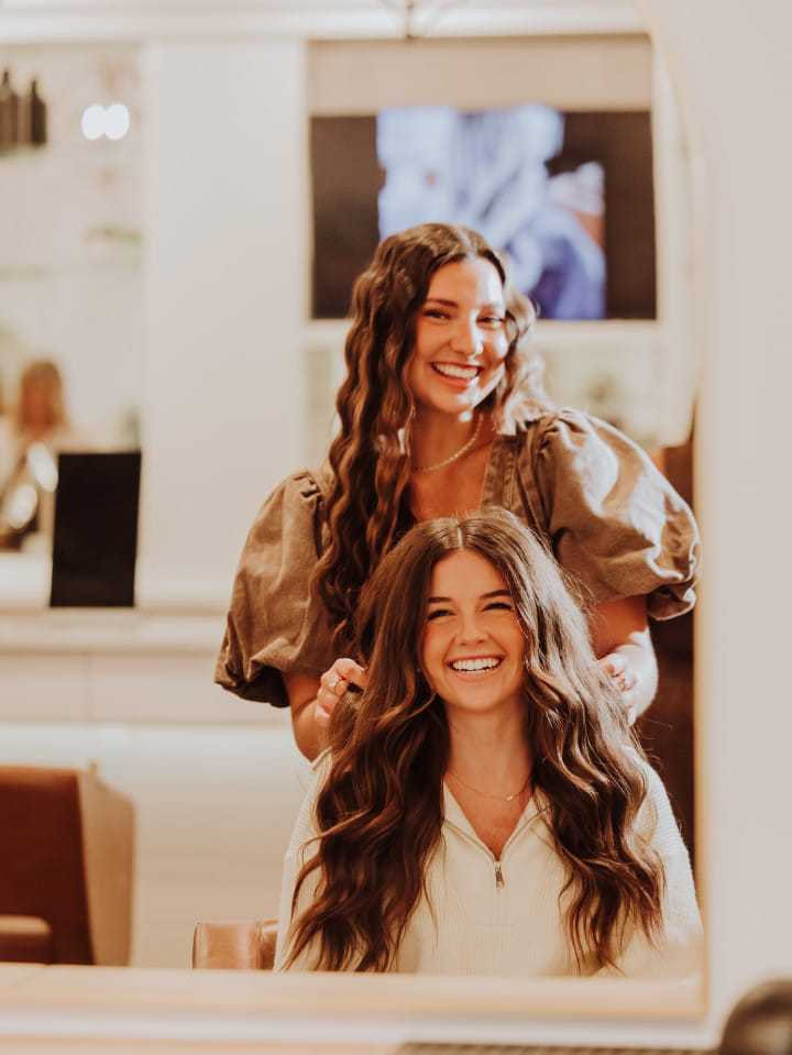Two women with long hair smiling in a salon, one styling the other's hair.