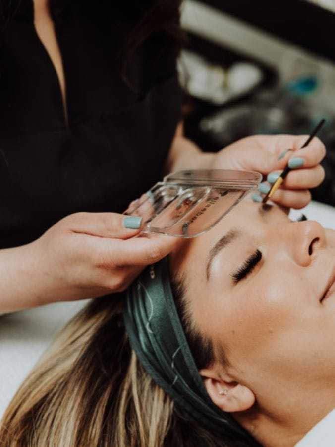 Person receiving eyebrow shaping with a stencil and brush at a salon.